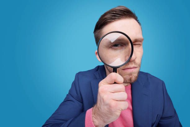 Man wearing a pink shirt and blue blazer looking through a magnifying glass. 