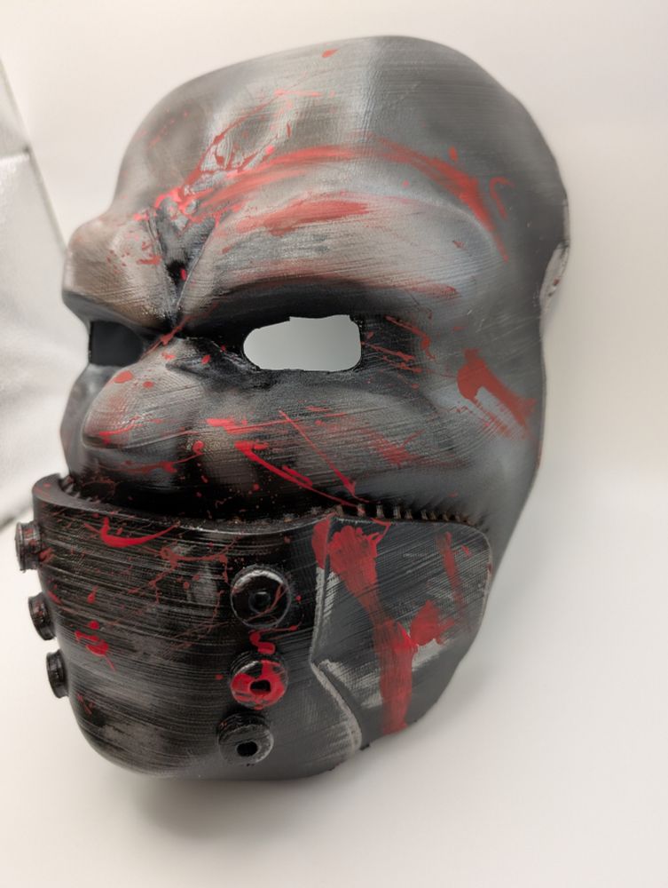 Grey mask of angry looking face with mouth covered by metallic looking apparatus hooked into it. Mask is covered by splats of red and sneaked in some places simulating somewhat fresh looking blood smears.