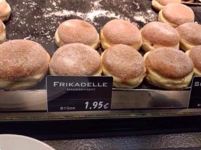 Im Vordergrund ein Schild, auf dem „Frikadelle“ steht. In der Auslage dann Berliner/Krapfen.