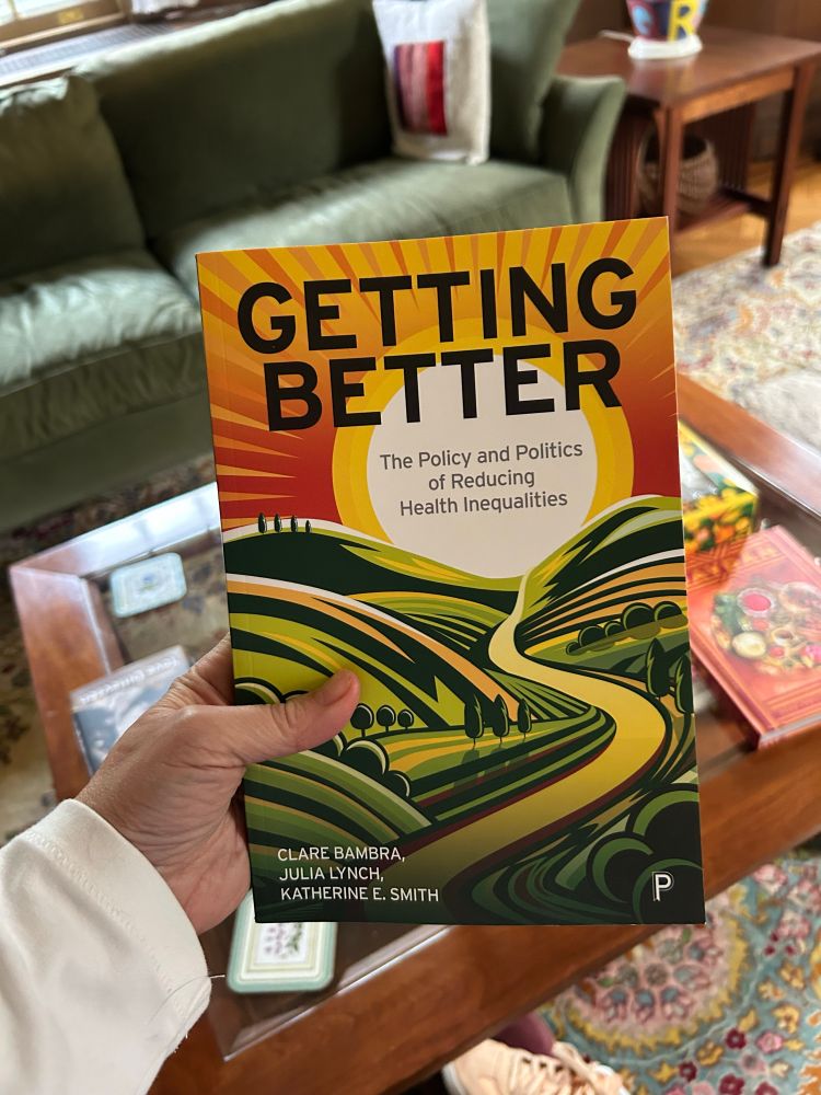 Photo of an author’s hand holding a paperback book.  The book is Getting Better: The Policy and Politics of Reducing Health Inequalities, by Clare Bambra, Julia Lynch and Katherine E. Smith. The cover image is of a rising sun and sunlit uplands, in colors of yellow, orange and green. 