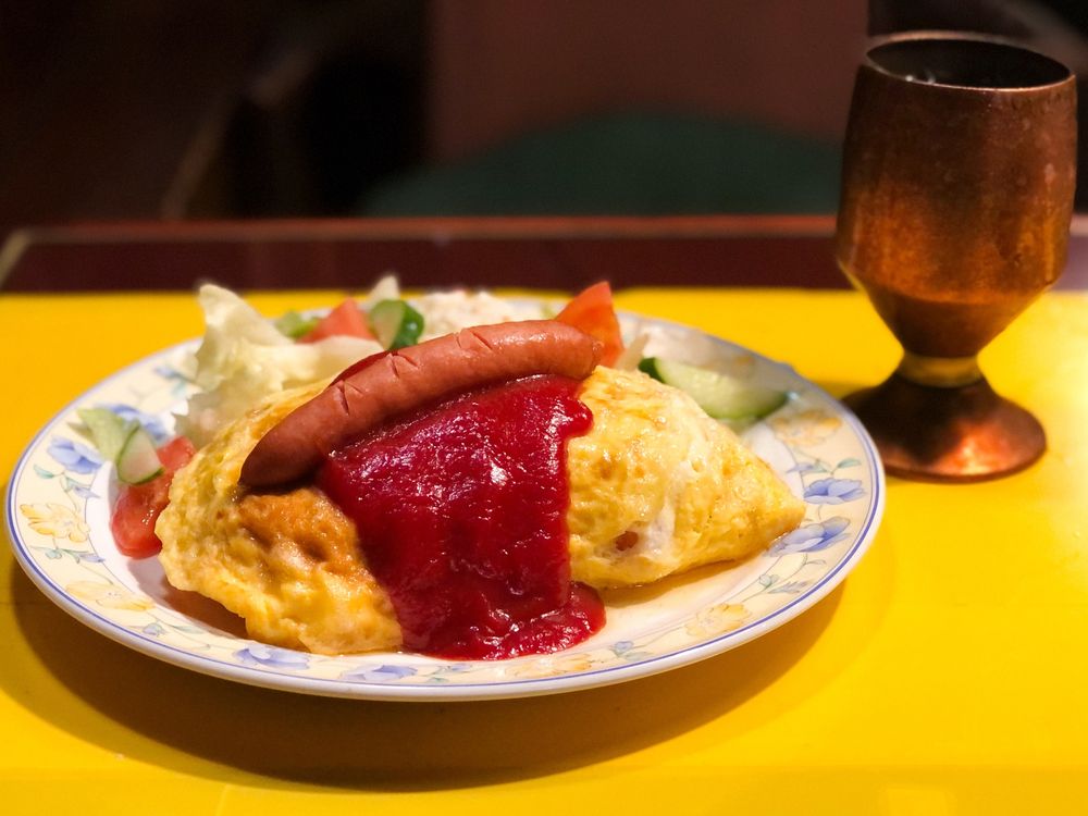 ケチャップがたっぷりとかかったオムライスにソーセージが添えられた一皿。彩り豊かなサラダが添えられ、レトロなカフェの温かみを感じさせる盛り付け。