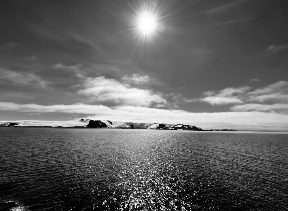 A black-and-white photograph of the first glimpse of Antarctica from the sea, with a brooding sky and the light of the overhead sun reflected in the water. 