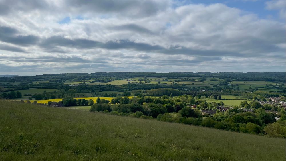 A scene from a hilltop with fields and trees below. Some of the fields are bright yellow with rape seed flowers. The sky is cloudy with patches of blue.