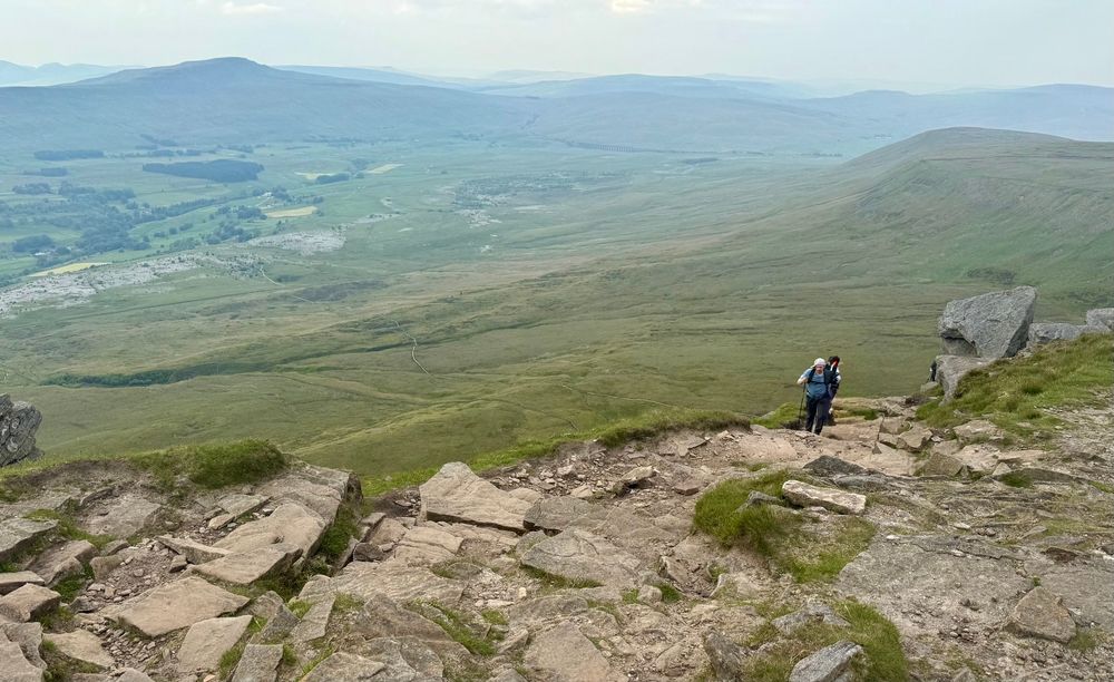 Beautiful view of the Yorkshire dales from the top of Ingleborough. A trekker is just coming up to the rocky summit up a steep path. 