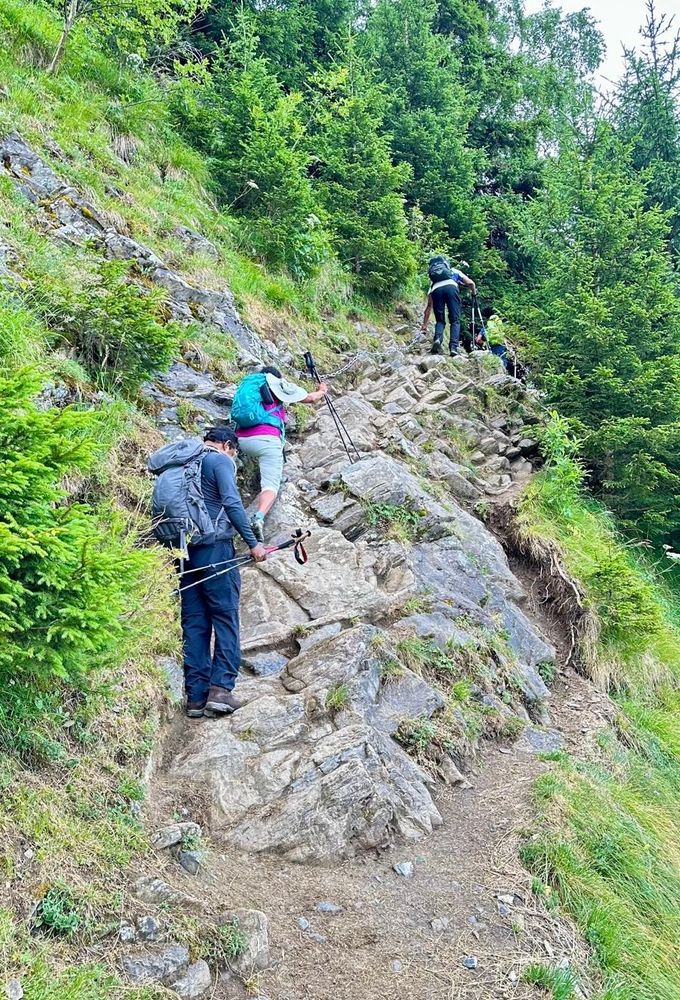 Trekkers navigating a tricky section of the Tour de Mont Blanc trek involving scrambling over rocks, with a sheer drop to one side. 