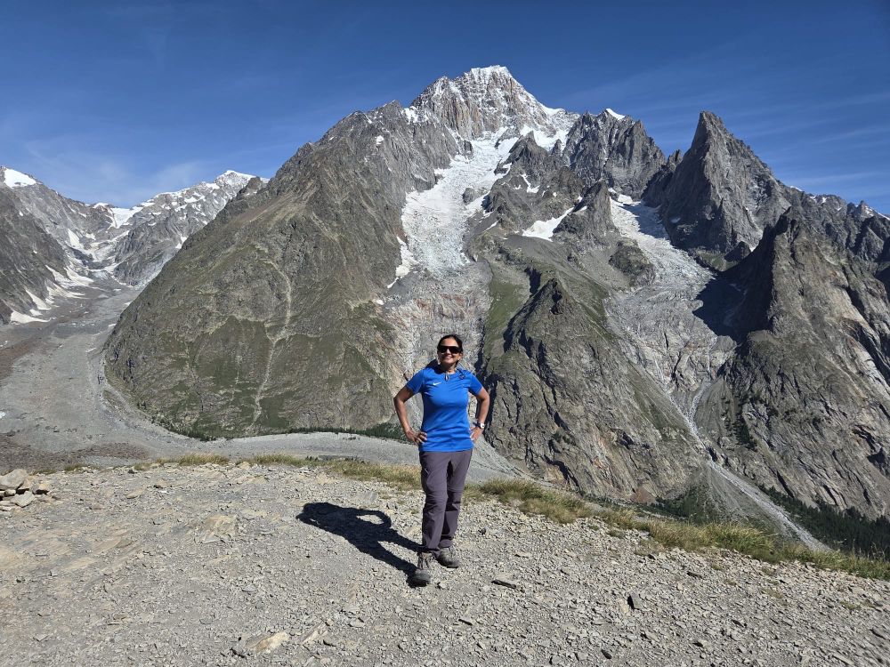 Photo of me in the Alps near Courmayeur. Greyish brown mountain peaks with streaks of snow are seen in the background. 