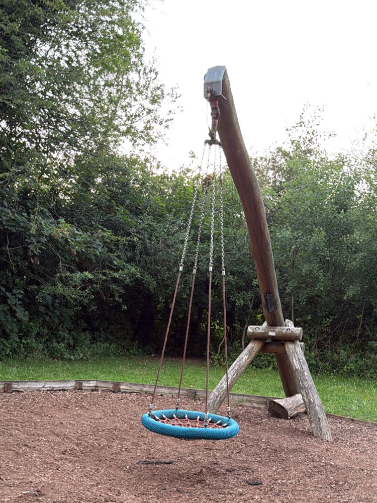 A round bucket swing attached to an inverted Y shaped wooden support, in a children’s play area with wood chippings on the floor and trees surrounding the swing.