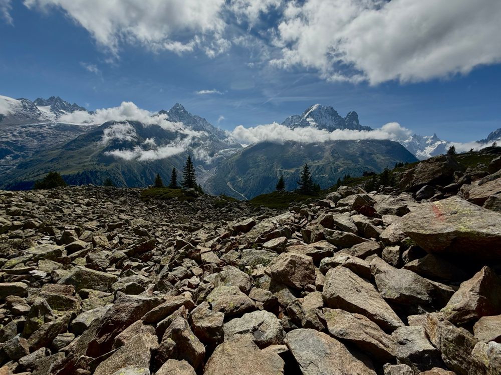 A photo of rocks with sharp edges in the foreground with no obvious walking path visible to move forward on, with bluish green mountain peaks wreathed in clouds seen in the distance. The sky is blue with more clouds. 