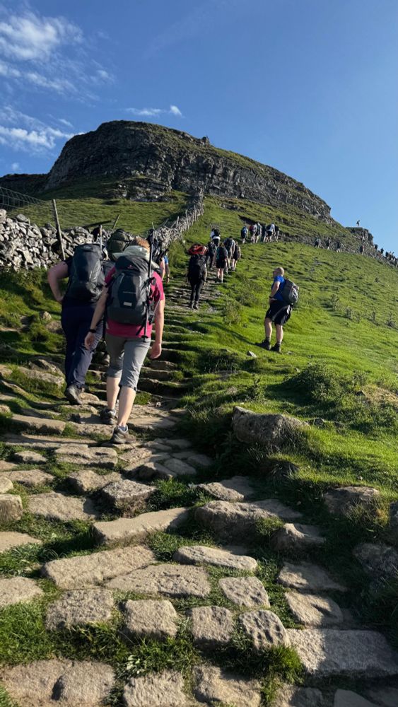 Photo of people with backpacks climbing a tall hill with a rocky summit. It’s sunny with a bright blue sky. 