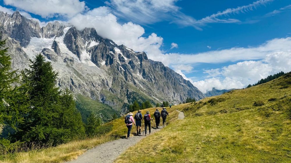 A photo of trekkers walking alongside a panorama of the Alps. The sky is blue and streaked with clouds. 