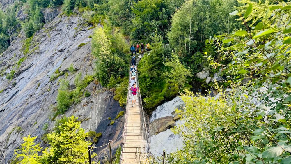 A suspension bridge with walkers crossing it. A stream can be seen among rocks far below, with greenery in foreground. 