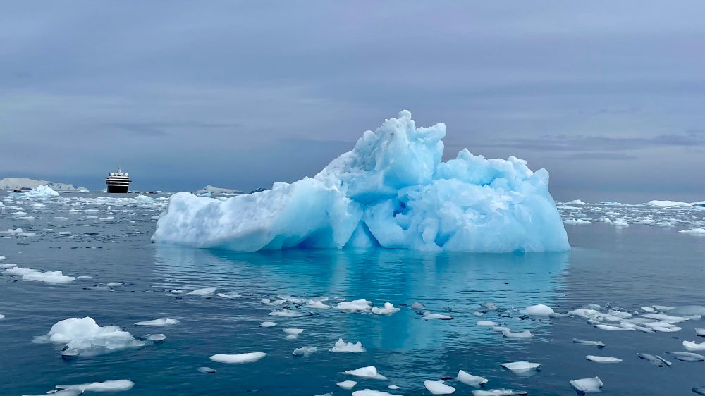 An image of a large bluish white iceberg floating in the sea in Antarctica with smaller fragments of ice surrounding it. A tiny ship is visible on the horizon. The part of the iceberg which is below water is seen clearly. 