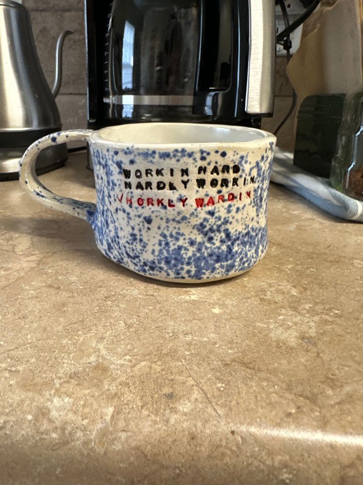 A wonky mug with blue and white speckled glaze. It has the words on it:
HARDLY WORKIN
WORKIN HARD
✔️HORDLY WORKIN