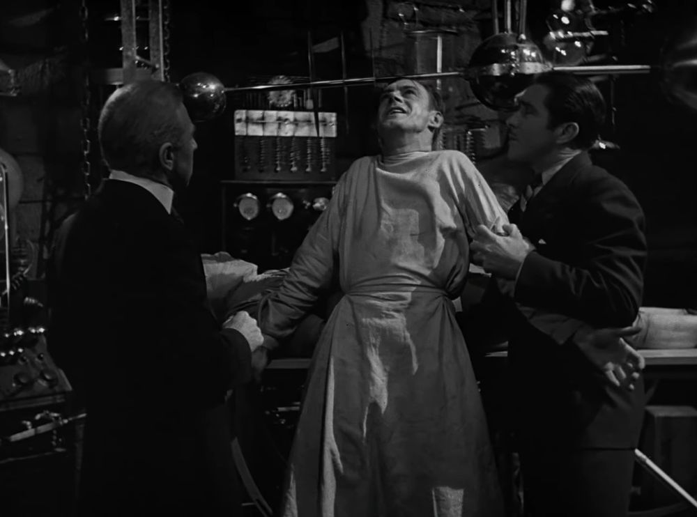 Frankenstein in rapture staring upwards now that his creation (visible behind him) is alive. His friends Moritz and Waldman are either side of him, shocked at what is happening.