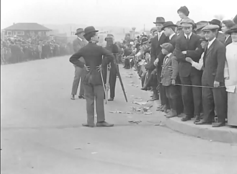 From behind this time, Chaplin striking a pose with hands on his hips. People line up either side.