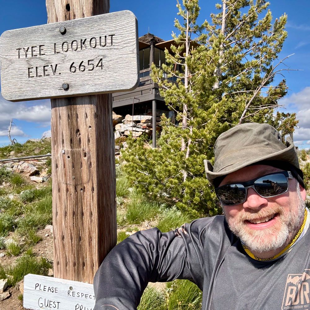 sign post of Mount Taiye with the elevation 6654 with a bearded motorcycle rider wearing sunglasses and hat, leaning against the post. Taken from the top of the mountain 