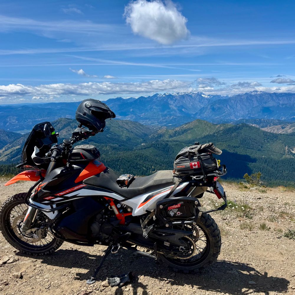 Picture of a KTM Adventure 890 motorcycle on its kickstand at the top of a mountain. Helmet is resting on the handlebars. Mountain ranges in the background.