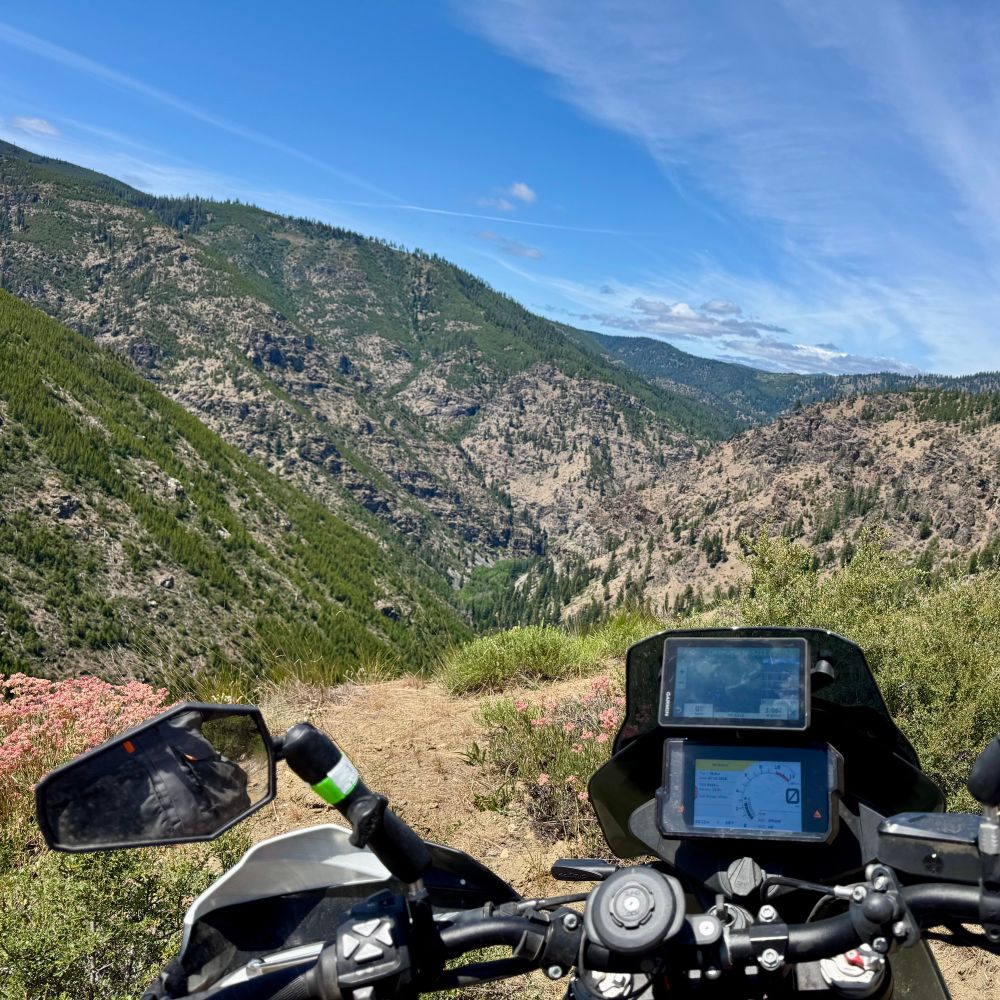 View of a mountain range in the valley from the top of the mountain with the handlebars and dash of a motorcycle in the foreground
