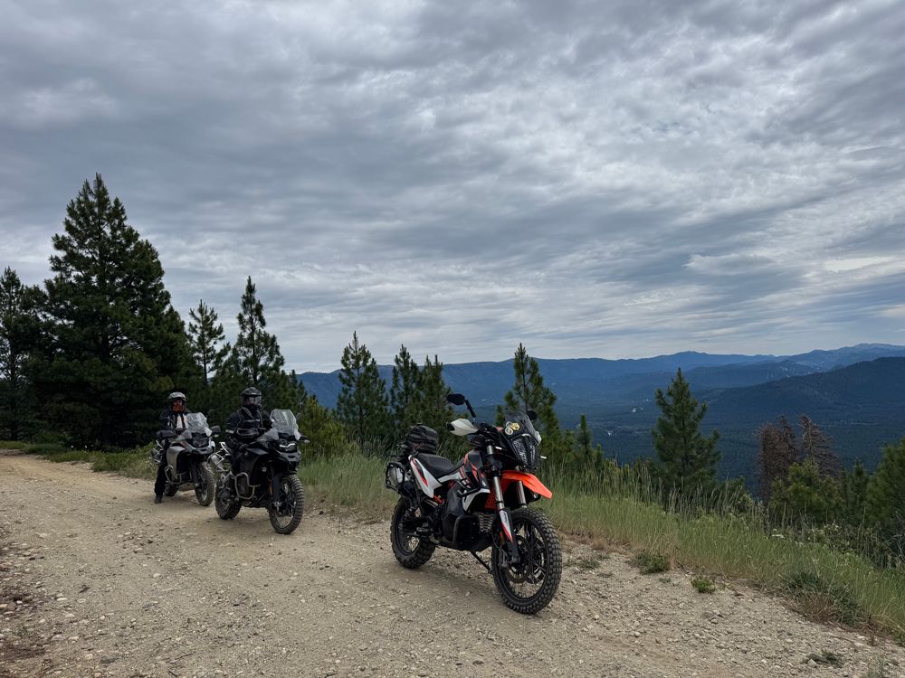 pictures of three motorcycles on a dirt road at the top of the mountain on their kick stands, with a valley down below