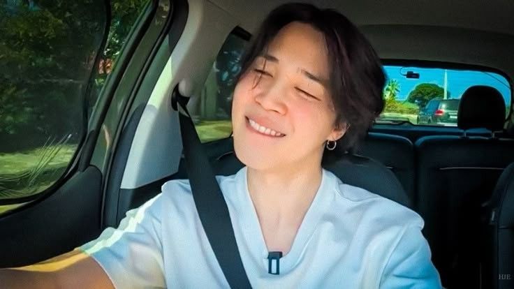 Jimin sitting inside the car, wearing a white t-shirt, with a happy smile on his face. AYS Jeju