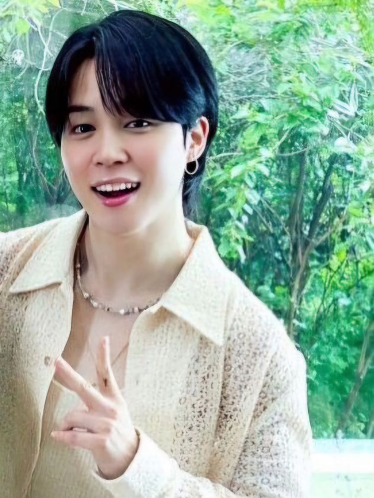 Jimin smiling at the camera. He is wearing a cream coloured opened-buttoned long sleeve shirt.