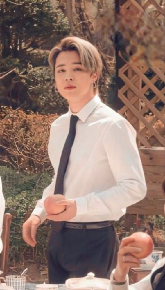 Jimin in a white long sleeved shirt with black tie and black trousers. He is also holding an apple.