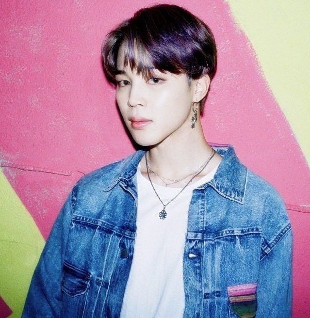 Jimin wearing a white shirt under a denim jacket. He is standing behind a colourful wall. Dynamite MV
