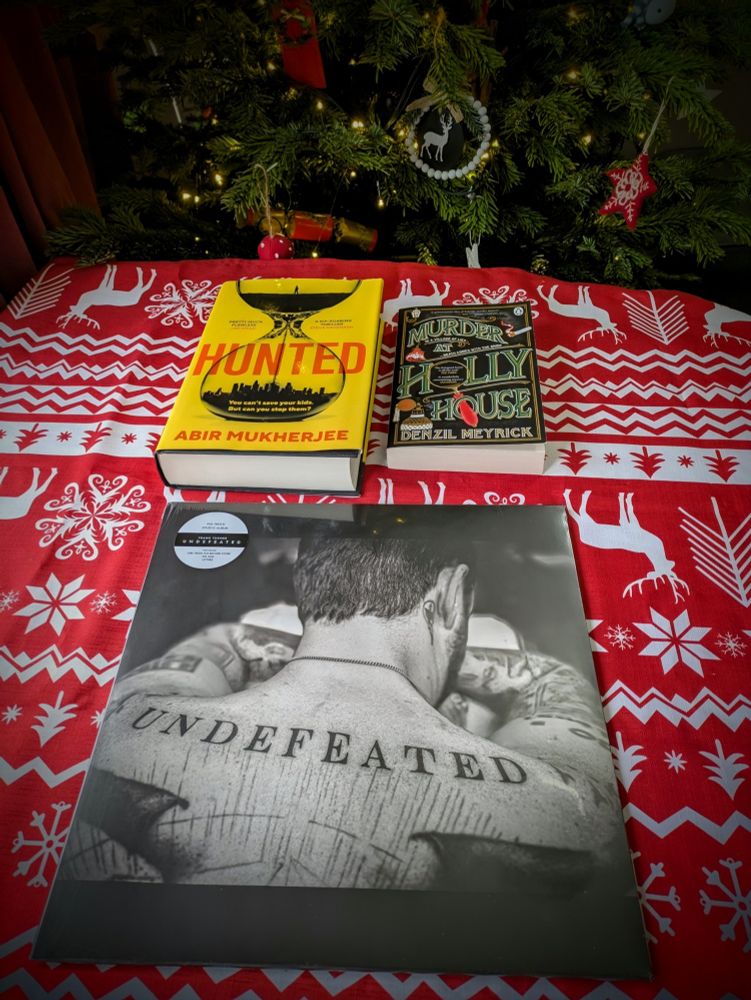 Image shows two books – Hunted by Abir Mukherjee and Murder at Holly House by Denzil Meyrick – and a vinyl LP record, Undefeated by Frank Turner, on a red and white Christmas tablecloth. Part of a lit Christmas tree is visible in the background.