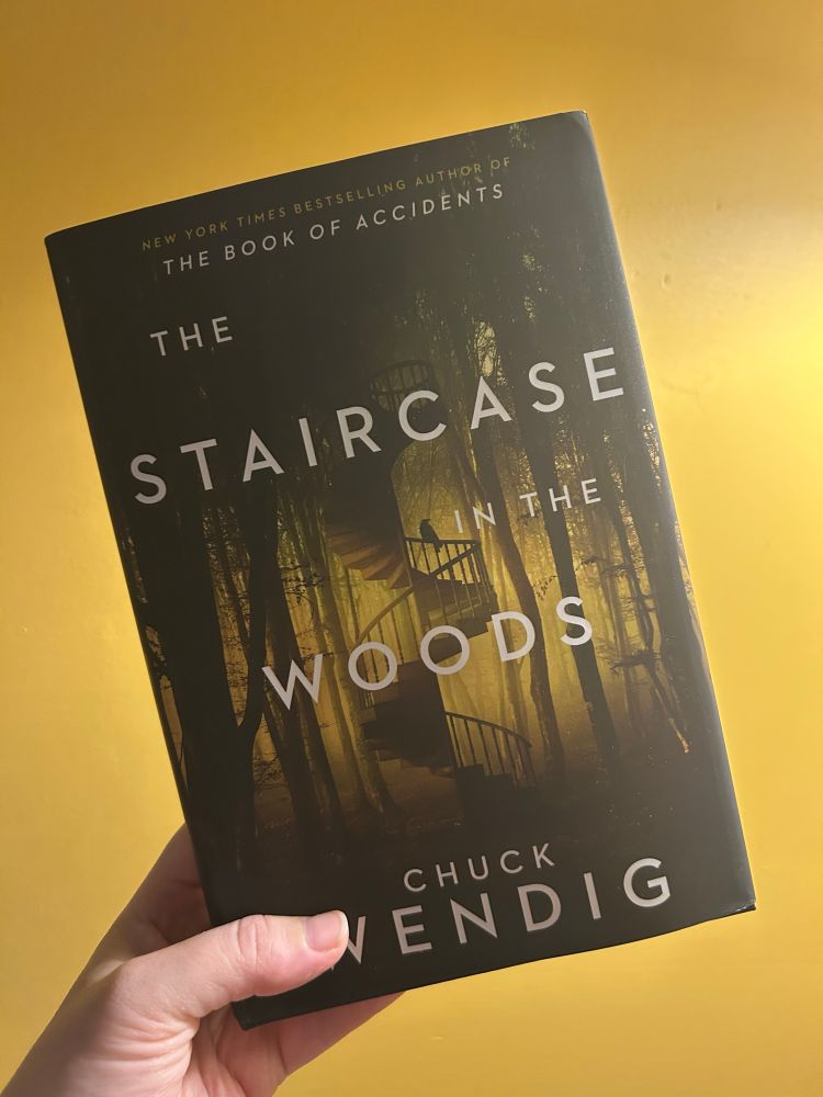 A hand holding a hardcover book called Staircase in the Woods by Chuck Wendig, against a yellow wall. 
