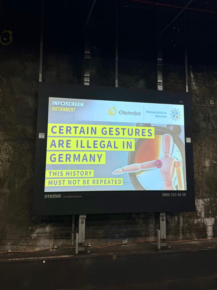 info screen bahnhof münchen mit dem text: certain gestures are illegal in germany. this history must not be repeated. daneben abgebildet: zeichenpuppe, die den rechten arm unter schulterhöhe ausgestreckt hat.