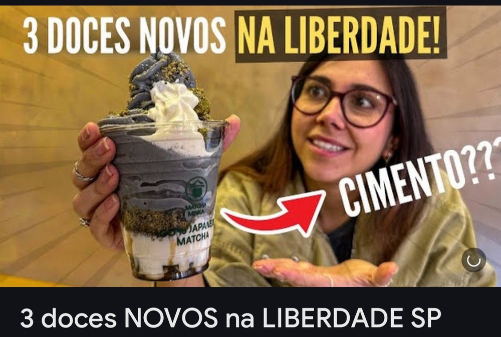 Print de miniatura de vídeos, mostra uma mulher de cabelo preto com óculos de grau e armação escura segurando um sorvete cinza com branco e olhando para ele com surpresa. Em cima tem o texto "3 doces novos na liberdade!" E uma seta aponta pata o sorvete com o texto "cimento??"