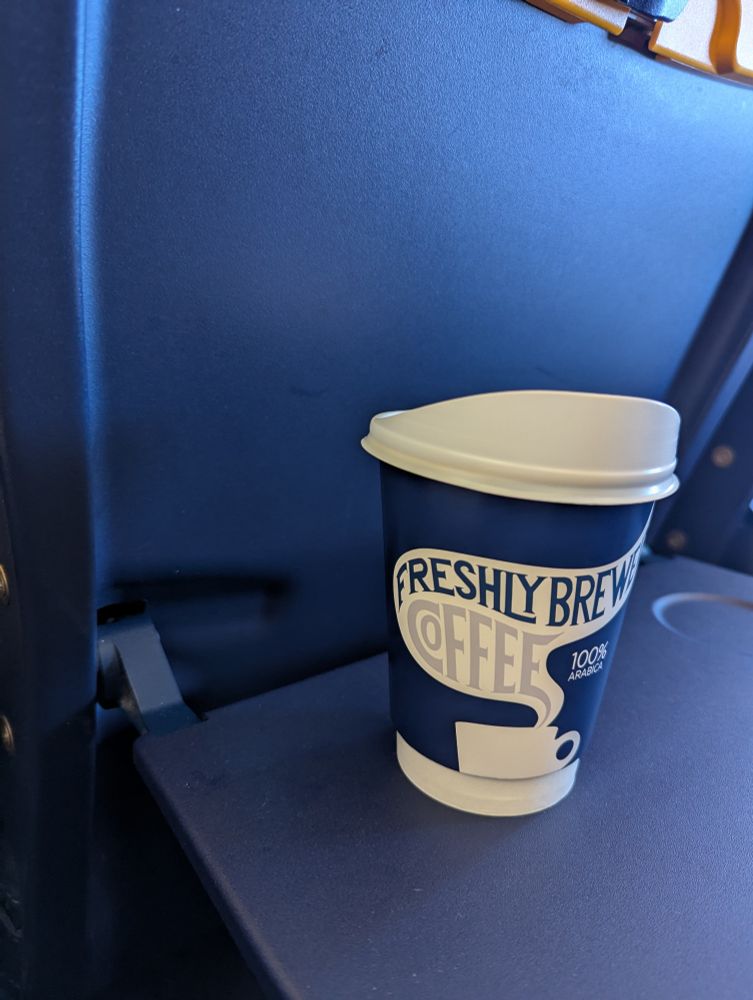 A cup of coffee in a takeaway mug, sitting on an airplane tray table. Words say "Freshly brewed coffee"