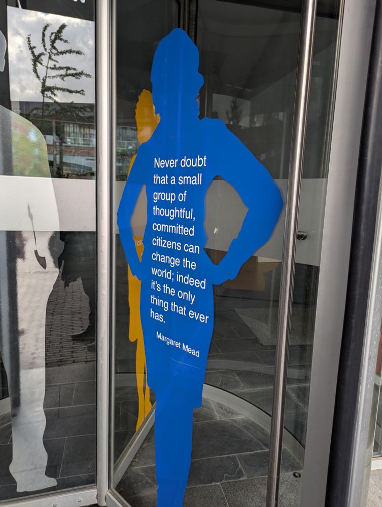A picture of some revolving doors with prints on the clear glass. One print depicts a figure, with the words "Never doubt that a small group of thoughtful, committed citizens can change the world; indeed it's the only thing that ever has. Margaret Mead"