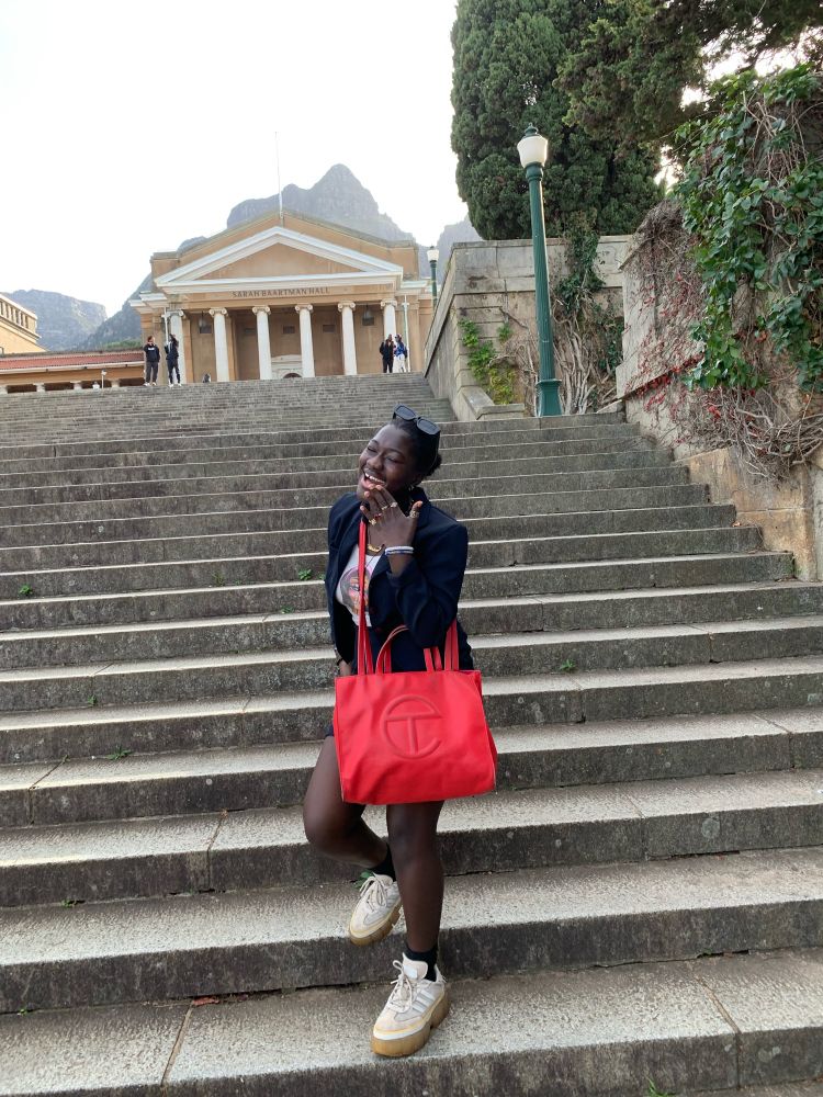 A dark-skinned baddie stands at the bottom of a long staircase. They have on a medium sized red Telfar bag and have been captured mid-laugh, one hand raised to cover their mouth. In the horizon is a beautiful mountain and a tall building with several large white columns. Along the staircase is some beautiful green foliage that leads up to the building.