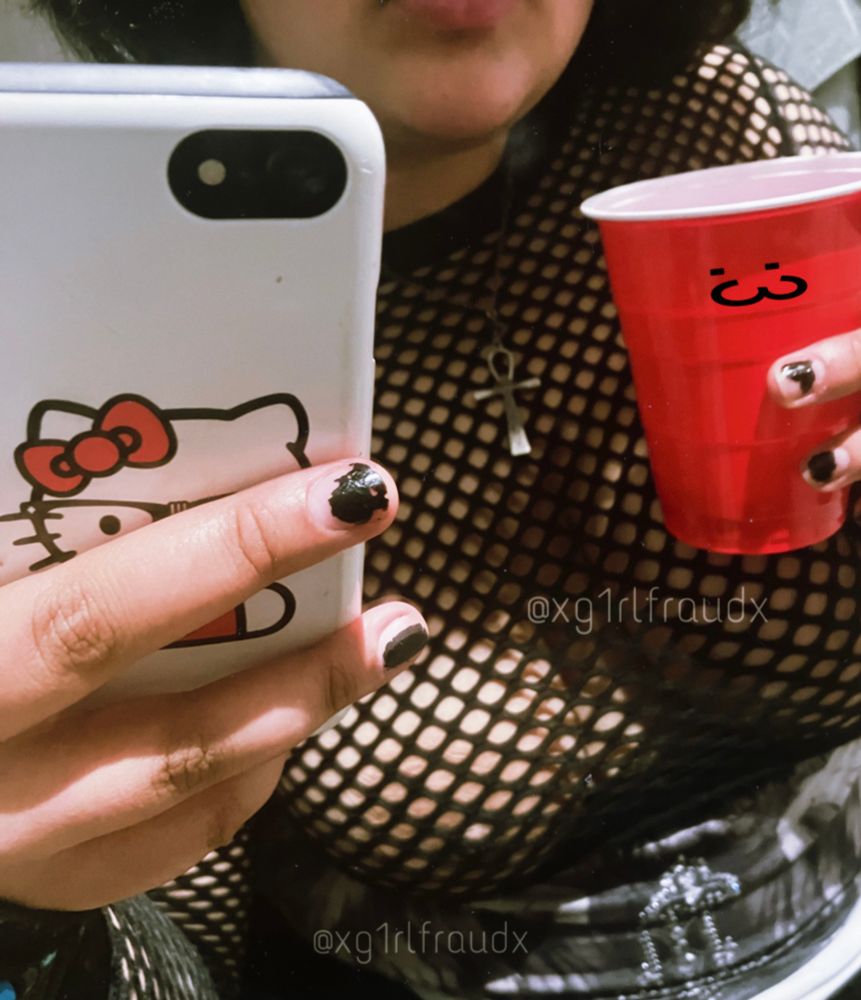 me taking a mirror selfie with my white hello kitty phone case in frame close to the camera and a red solo cup in my other hand with a colon 3 smiley face sticker on it. i’m wearing a fishnet top that you can see my tits through, and i have chipped, black nail polish on my fingers. my face is just out of frame