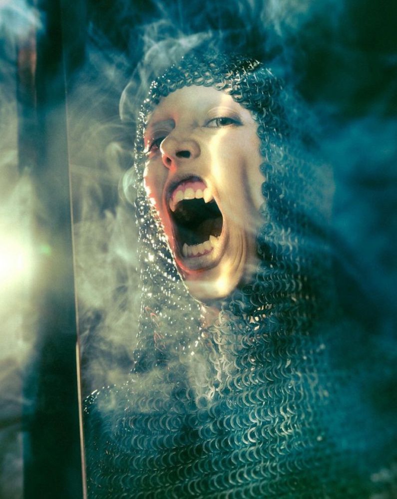 Cold colors. A portrait of the knight from a low angle, clouds of smoke around her chainmail covered head, her mouth open, she is baring her long, sharp fangs. 