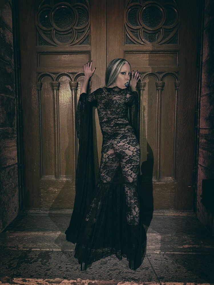 Ophelia, the goth with a heart that is looking for legendary romance, pushing against the heavy doors of a cathedral. She is wearing a black see through lace gown with nothing underneath, letting you almost see her entire petite body. She is looking back at you over her shoulder, with an expression that feels like an invitation.