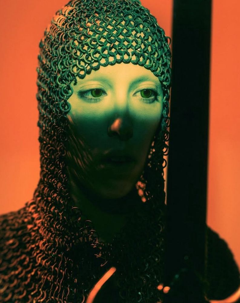 Strong contrasts of green against orange. A closeup portrait of a Knight in chainmail, looking at her sword, deep in thought, her eyes are covered in light, the lower half of her face lies in the shadow.