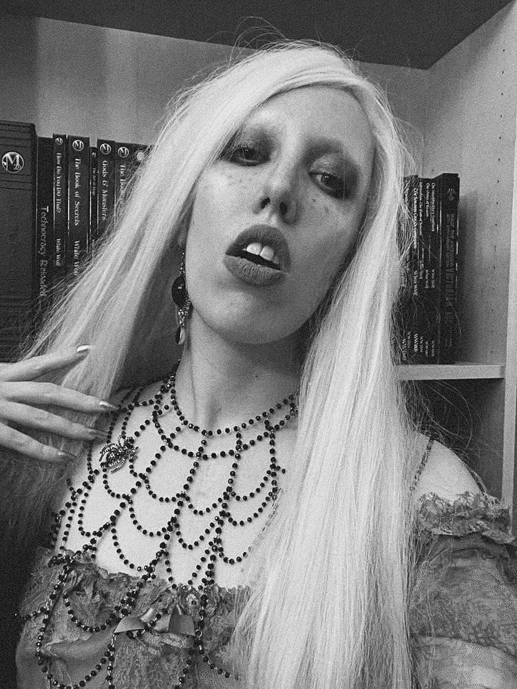A black and white potraitof Ophelia. Her Long white hair flows over her shoulders, her lips are parted. Her necklace is in the Share of a spider‘s web made out of black pearls.