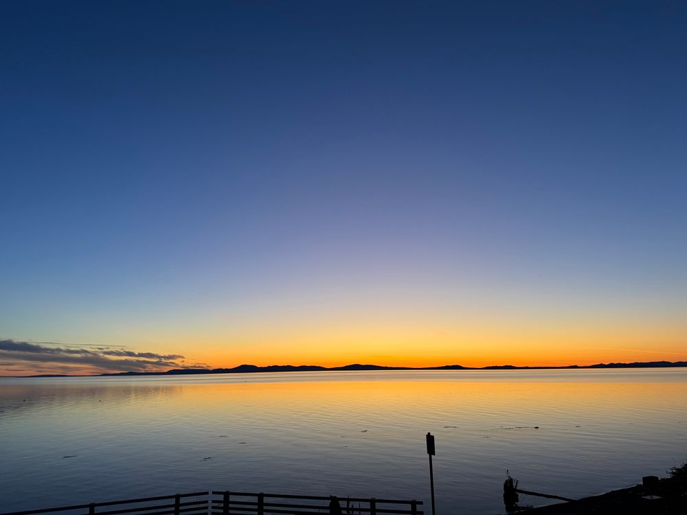 A twilight photo of Kotzebue Sound. The  water is still as glass and the distant hills across the sound are framed in yellow orange glow accentuated by smoke from tundra fires in northern Alaska. The sky is almost gloaming blue.