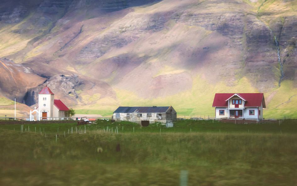 A small church, a home and what looks like a barn are seen in the distance, a field before them and a hill behind 