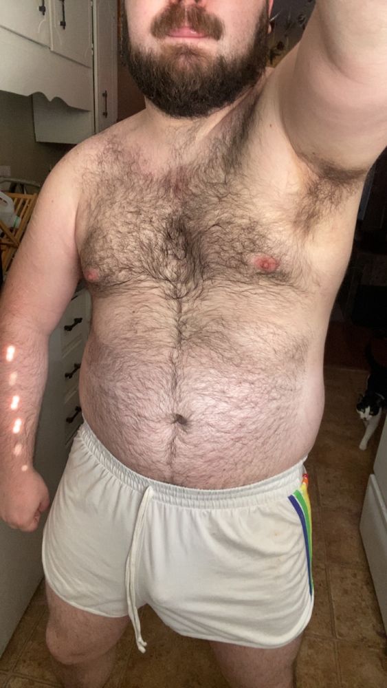 A selfie of me standing in my kitchen. I’m shirtless and sweaty, wearing short white gym shorts with VPL. Our cat, a black and white tuxedo named Titus, strolls in stage right.