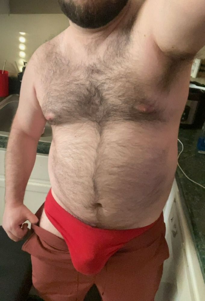 A top down selfie of me standing in my kitchen, partially undressed. My hairy chest and stomach are in frame as I pull down my brown pants to show my big soft bulge in a pair of red underwear. The countertop behind me has various kitchen items.