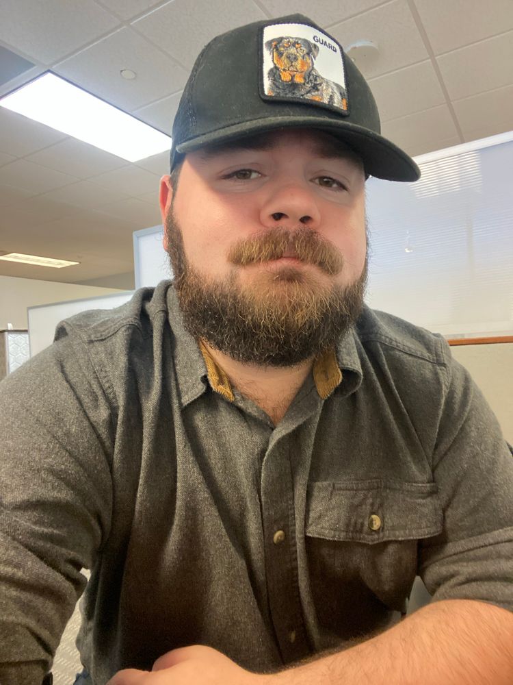 A selfie of me at my desk, same outfit as the previous image, wearing my black cap with an embroidered image of a Rottweiler on it.