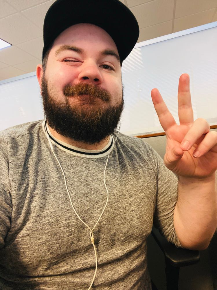 A picture of me in my cubicle, winking and holding up the peace sign. I’m wearing a grey shirt, sleeves rolled up to the elbows, as well as a black cap.