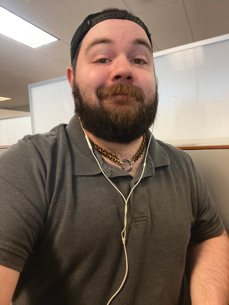 A picture of me in my cubicle, I’m wearing a grey polo and my pup collar, as well as a pair of EarPods and a backwards cap. I’m smirking at the camera.