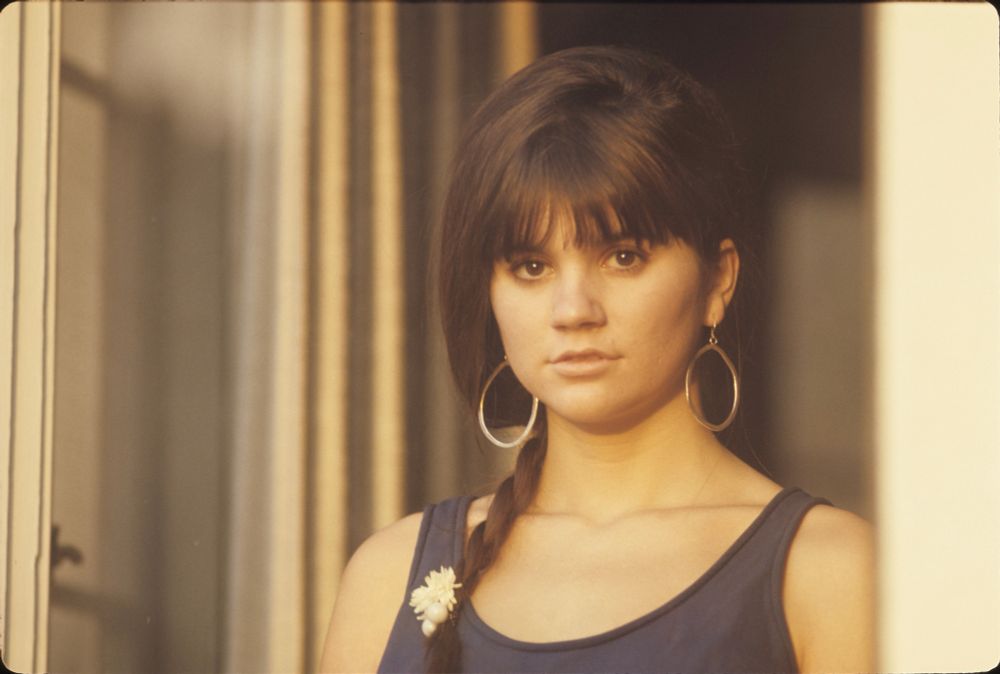 A color photo of a young Linda Ronstadt. She’s standing on the other side of what appears to be a sliding door. Behind her, curtains can be hanging in the background. The entire scene is warm and sunlit. Linda has her hair meticulously crafted into a braided ponytail, her loose hair hanging at the sides like a bob. A flower, possibly a daisy, sits at the tip of the braid that crosses her neck and goes down her décolletage. She’s wearing a navy blue top with a low swoop connecting the the tank top-like straps. She looks forward with a soft, but striking neutral expression, her face framed by golden hoop earrings.