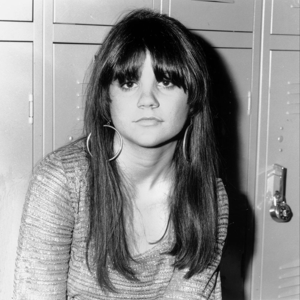 A young Linda Ronstadt, picture dated 1968, age 21. She is standing in front of a row of high school lockers and looks forward with a neutral, but confident expression. Hoop earrings peek out from either side of her long hair, the gently parted bangs obscuring her eyebrows. She is wearing what appears to be a longs sleeved sequined shirt with a low scoop collar.