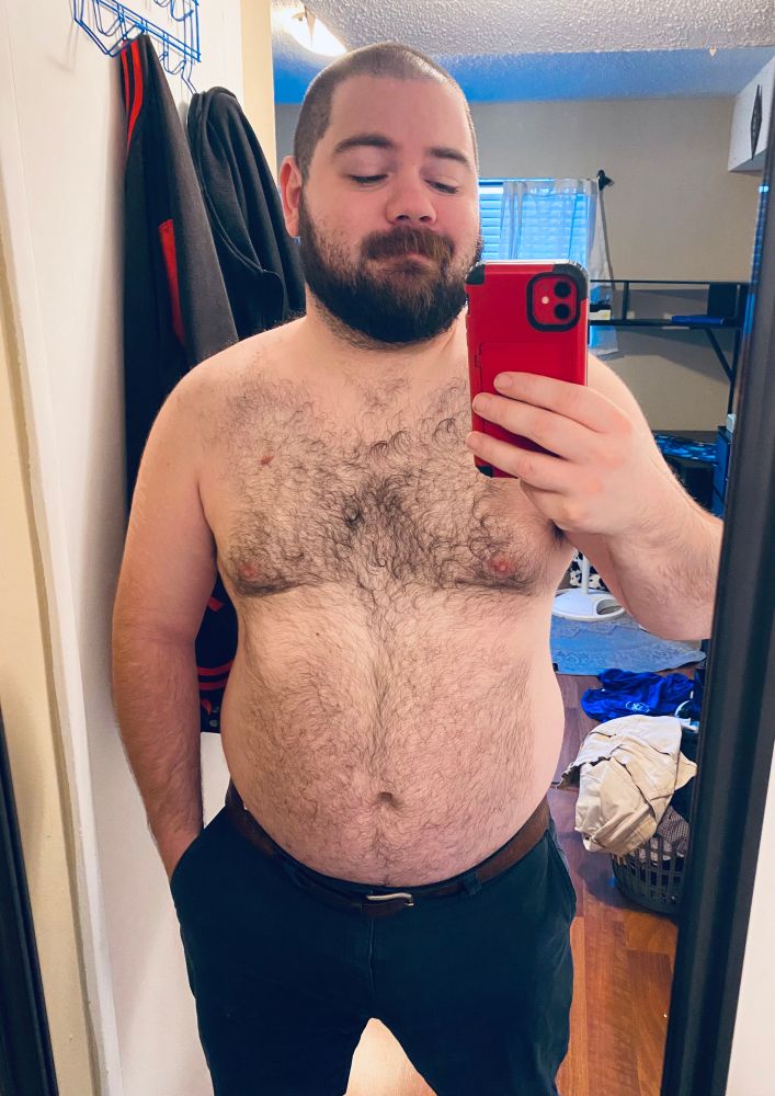 A selfie of me standing shirtless in front of a mirror, holding my red phone up to take the picture. I’m wearing navy blue chinos and a belt, and behind me is my bedroom— with a computer desk and laundry baskets visible in the background.