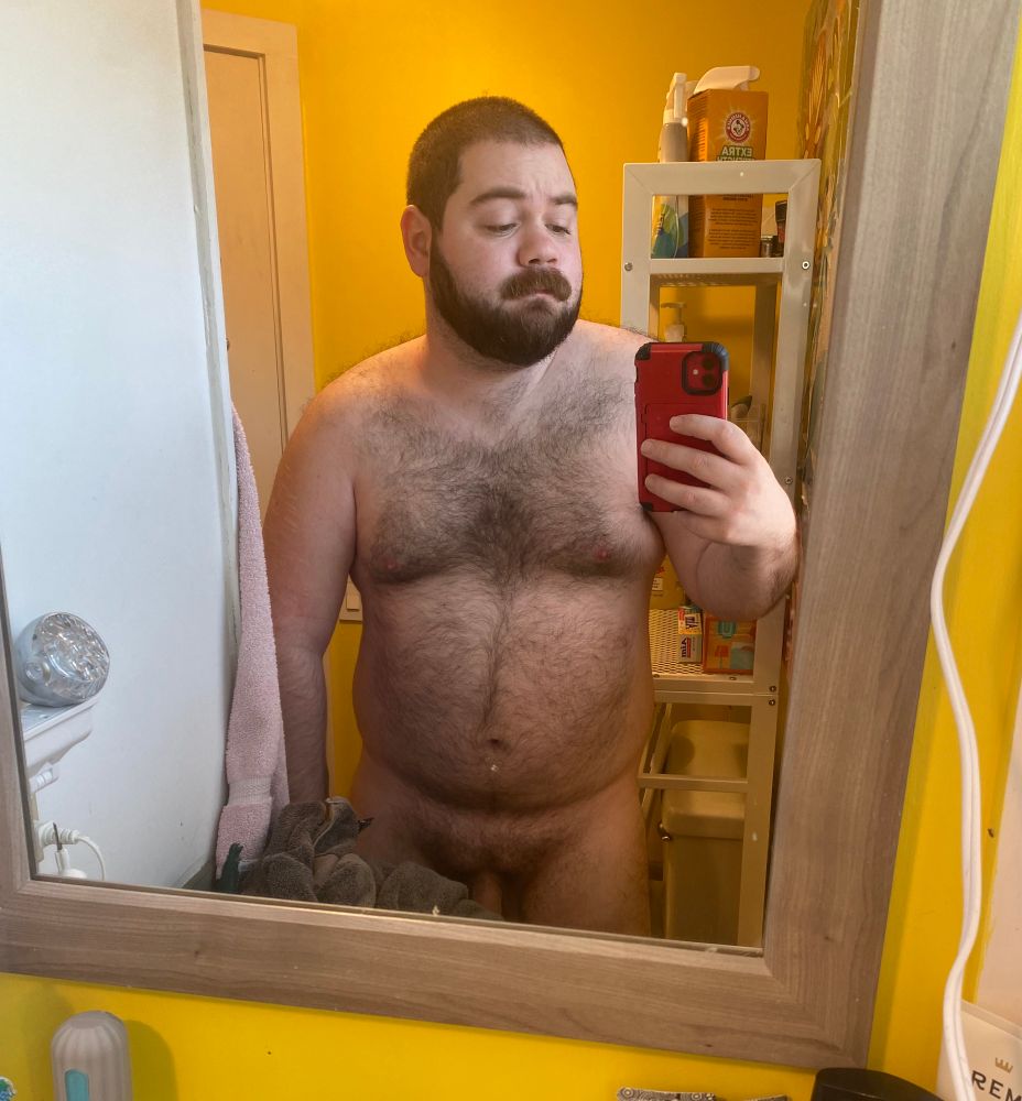 A mirror selfie of me standing in a yellow bathroom, holding up my red phone to take a pic. I’m nude, hairy, and beefy.